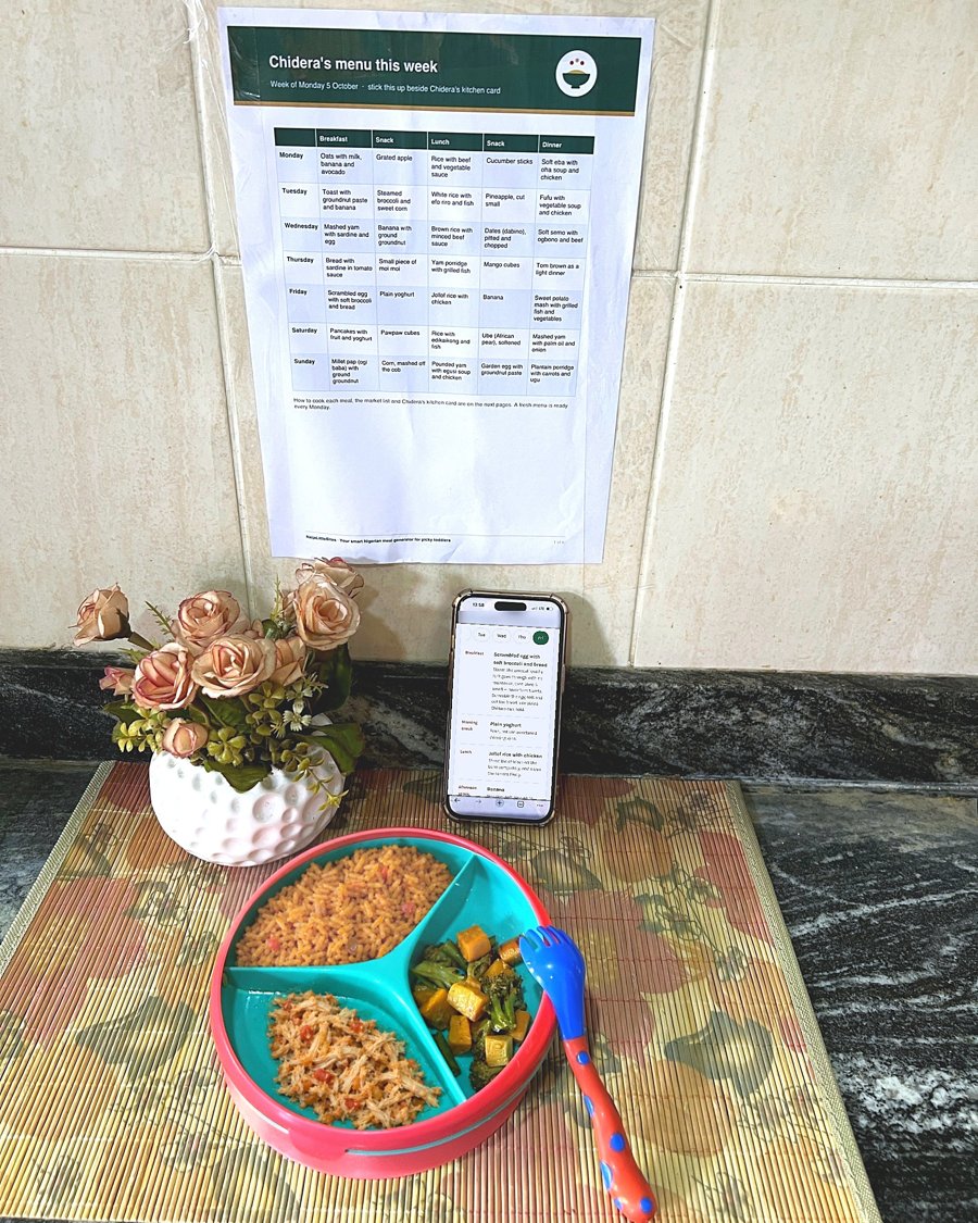 A phone showing the meal tool beside a printed weekly menu on a kitchen wall, with a toddler's plate of jollof rice